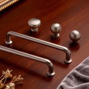 May include: A collection of brushed nickel cabinet hardware, including two bar pulls and three round knobs. The hardware is displayed on a dark wood surface, showcasing the metallic finish and simple design.