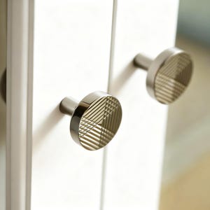 May include: Close-up of two silver cabinet knobs with a geometric, striped design on a white cabinet door. The knobs have a circular face and a cylindrical stem. The design is a modern, minimalist style.