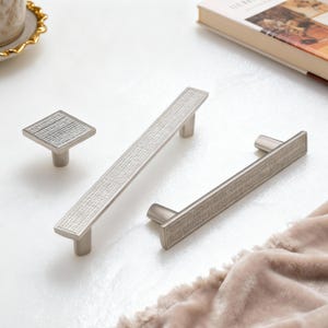 May include: A set of three silver cabinet hardware pieces: a square knob and two rectangular pulls. Each piece has a textured, cross-hatched design on the front. The hardware is displayed on a white surface with a soft, neutral-colored fabric.