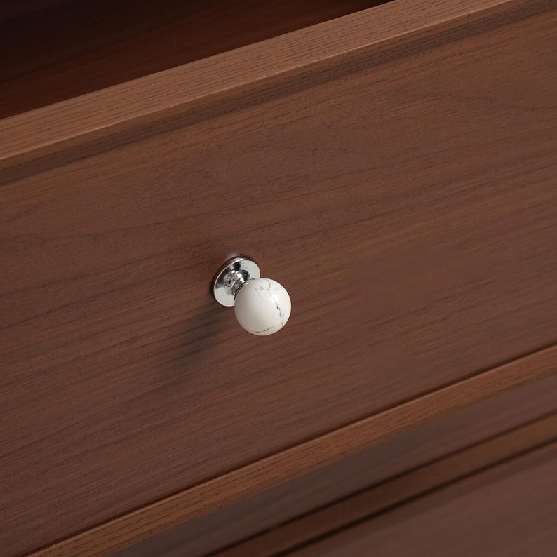 May include: A close-up of a wooden drawer with a white marble-effect knob. The knob has a chrome base and a spherical, patterned top. The drawer is made of dark brown wood with visible grain.