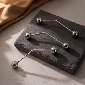Może przedstawiać: Trzy srebrzyste kolczyki typu industrial barbell, każdy składający się z wygiętego metalowego pręta z polerowaną kulką na obu końcach. Każda kulka jest wygrawerowana z małym symbolem gwiazdy, co zapewnia spójny wygląd wszystkich trzech elementów.