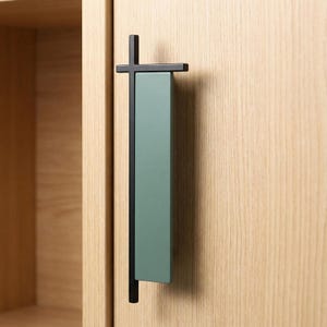 Puede incluir: Un tirador de armario moderno con una barra transversal negra y un panel vertical rectangular verde salvia. El tirador está montado en una puerta de armario de madera clara, mostrando un diseño minimalista.