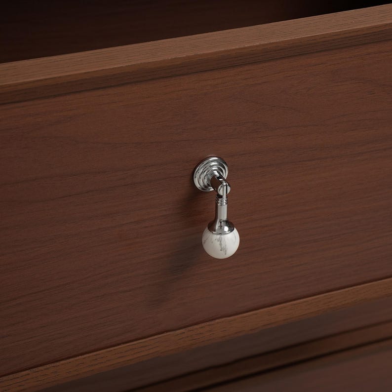 May include: A close-up of a wooden drawer with a silver and marble knob. The knob has a decorative design and a white and grey marble sphere at the end. The drawer is made of dark brown wood.