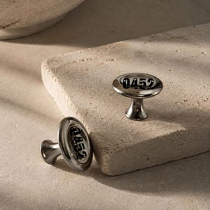 May include: Two polished dark metallic cabinet knobs with a smooth, rounded, mushroom-shaped top. Each knob features the black printed number 0452 centred on the top surface. The knobs have a short, tapered stem and a reflective, glossy finish.