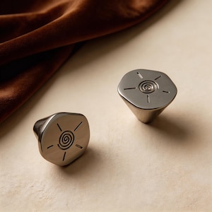 Puede incluir: Dos tiradores de armario de níquel cepillado con un diseño de sol. Los tiradores tienen una forma irregular y una base cónica. El diseño del sol presenta una espiral central con líneas radiales. Los tiradores se muestran sobre una superficie neutra.