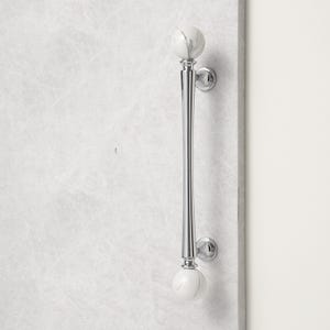 May include: A polished chrome door pull with white marble sphere accents. The long, slender handle features a tapered design and is mounted on a light-coloured surface. The door pull is a decorative hardware element.