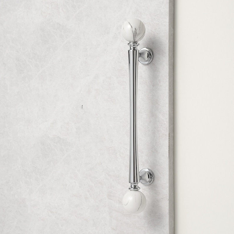 May include: A polished chrome door pull with white marble sphere accents. The long, slender handle features a tapered design and is mounted on a light-coloured surface. The door pull is a decorative hardware element.