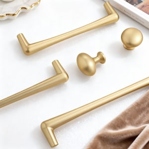 May include: A selection of gold-coloured cabinet hardware, including bar pulls and knobs. The hardware has a matte finish and a modern design. The pulls have an L-shaped design on each end. The knobs are round and oval-shaped.