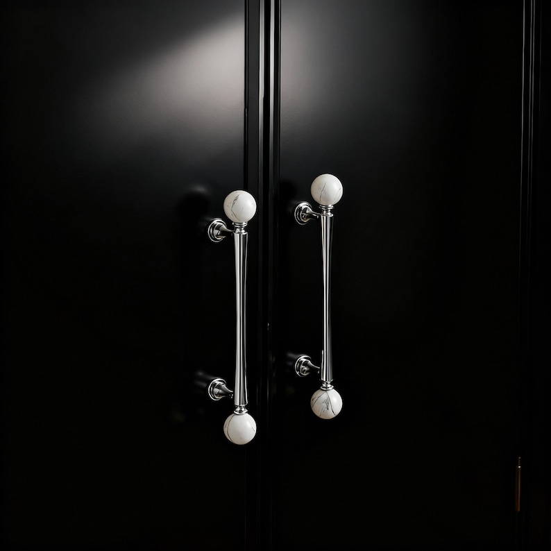 May include: Two cabinet door handles with a polished chrome finish and white marble sphere accents. The handles are long and slender, with a decorative design. The cabinet doors are black.