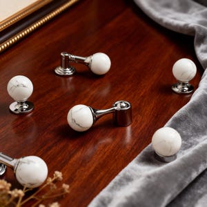 May include: Cabinet knobs and pulls with a white marble effect and grey veining. The knobs are mounted on a polished silver base. The pulls have a similar marble sphere with a curved silver arm, all set on a wooden surface.