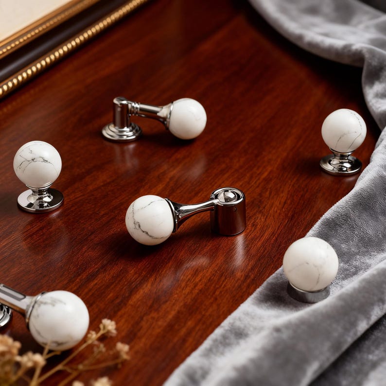May include: Cabinet knobs and pulls with a white marble effect and grey veining. The knobs are mounted on a polished silver base. The pulls have a similar marble sphere with a curved silver arm, all set on a wooden surface.