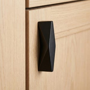 Könnte beinhalten: Ein schwarzer, geometrischer Schrankgriff an einer hellen Holzschranktür. Der Griff hat ein facettiertes Design und eine matte Oberfläche, die eine moderne Ästhetik bietet. Die Holzmaserung der Tür ist sichtbar.