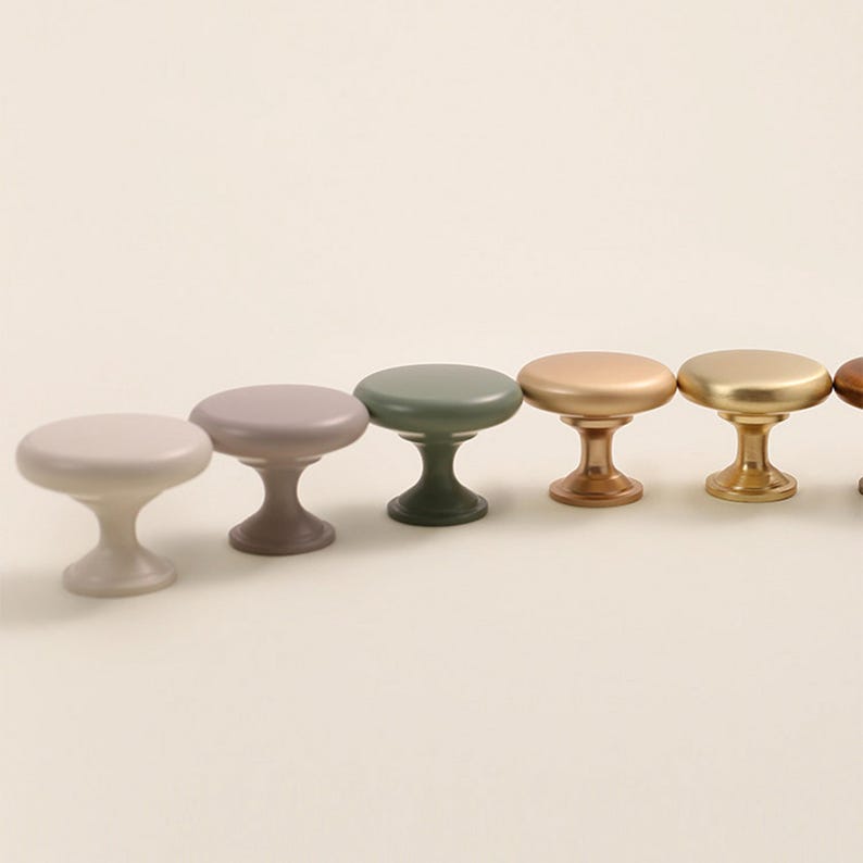 Pu&ograve; includere: Una fila di sei pomelli per mobili in vari colori, tra cui grigio chiaro, malva, verde, oro e bronzo. Ogni pomello ha una parte superiore arrotondata e una base a piedistallo, per mobili o decorazioni.