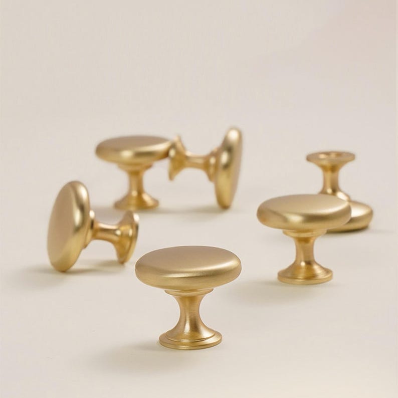 Pu&ograve; includere: Una collezione di cinque pomelli dorati per mobili. Ogni pomello ha una parte superiore piatta e arrotondata e una base a piedistallo. I pomelli sono disposti su una superficie chiara, mostrando il loro design e la finitura.