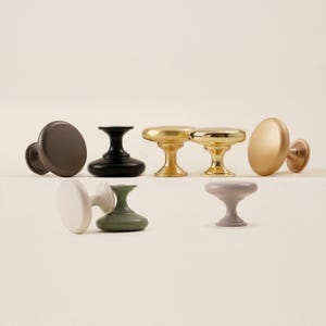 Pu&ograve; includere: Una collezione di pomelli per mobili in vari colori e finiture. I pomelli includono marrone opaco, nero, oro e tonalit&agrave; tenui di bianco, verde e grigio. Ogni pomello ha una testa rotonda o ovale e una base a piedistallo.