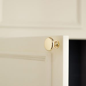 Pu&ograve; includere: Pomello per armadio dorato con una faccia rotonda e piatta e una base decorativa. Montato su un'anta bianca, con uno scorcio dell'interno. Questo hardware aggiunge un tocco di eleganza ai mobili.