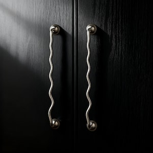 May include: Two silver-toned, wavy cabinet pulls are mounted on a dark, textured wooden surface. Each pull has a circular base and a decorative, undulating handle. The handles are symmetrical and create a modern aesthetic.
