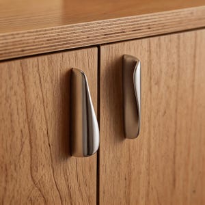 Könnte beinhalten: Nahaufnahme eines Holzschranks mit zwei Griffen aus gebürstetem Metall. Die Schranktüren bestehen aus hellfarbenem Holz mit sichtbarer Maserung. Die Griffe haben ein geschwungenes, ergonomisches Design.