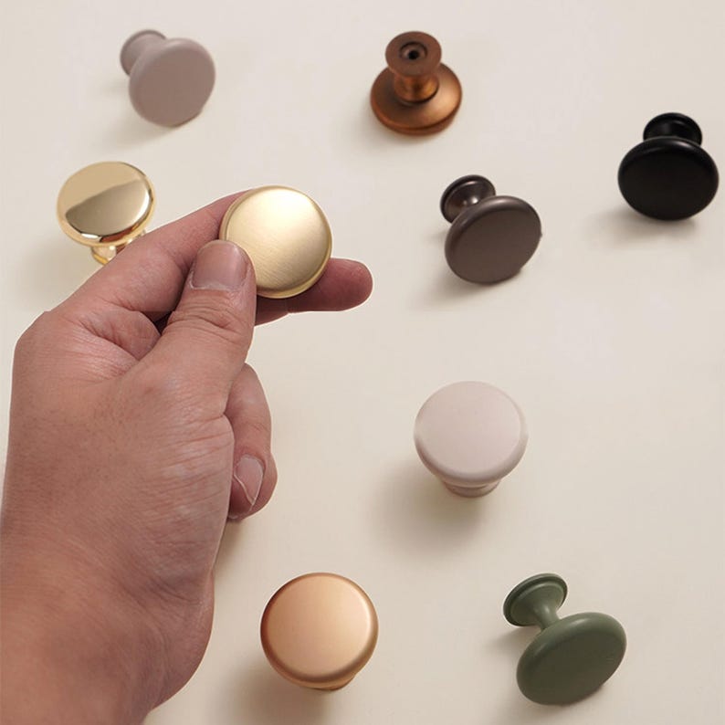 Pu&ograve; includere: Una selezione di pomelli per mobili in vari colori e finiture, tra cui oro, bronzo, nero e crema. Una mano tiene un pomello dorato, mostrando i diversi stili disponibili per l'arredamento della casa e il miglioramento dei mobili.