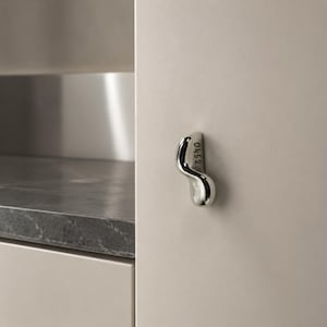 May include: A polished chrome cabinet handle with an organic, S-shaped silhouette mounted on a matte beige cabinet door. The side of the metallic handle features the engraved number 0452, providing a distinct detail against the smooth surface.