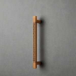 May include: A bronze-colored door pull with a rectangular design. The handle has a textured, grid-like pattern down the center and square ends. The hardware is mounted on a gray surface.