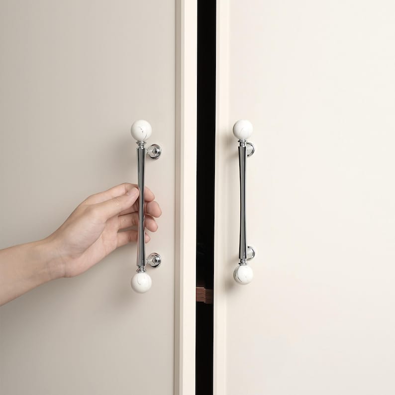 May include: Cabinet door handles with a polished chrome finish and white, marble-effect spheres. The handles are mounted on a light-coloured cabinet door. Each handle is approximately 20 cm in length.