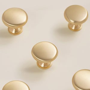 Pu&ograve; includere: Diversi pomelli per mobili color oro con finitura opaca sono disposti su uno sfondo chiaro. Ogni pomello ha una parte superiore arrotondata e una base cilindrica corta. I pomelli sono distanziati in modo uniforme, suggerendo un set.