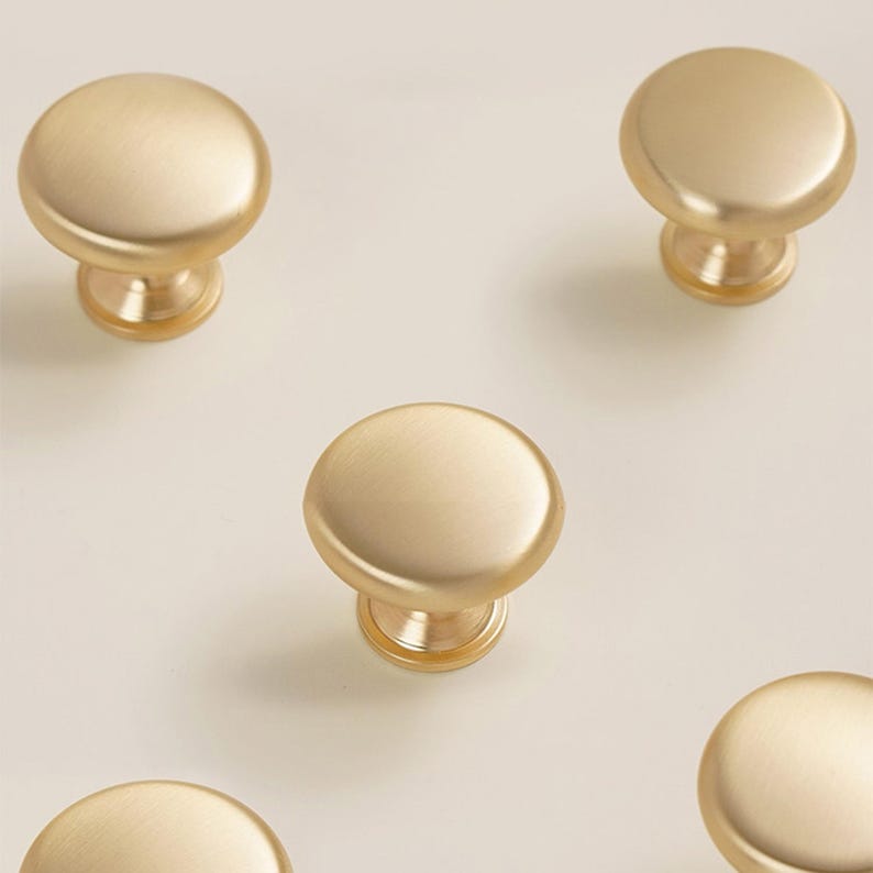 Pu&ograve; includere: Diversi pomelli per mobili color oro con finitura opaca sono disposti su uno sfondo chiaro. Ogni pomello ha una parte superiore arrotondata e una base cilindrica corta. I pomelli sono distanziati in modo uniforme, suggerendo un set.