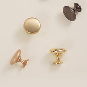 Pu&ograve; includere: Una collezione di pomelli per mobili in varie finiture metalliche. I pomelli includono colori oro, bronzo e rame. I pomelli hanno forme diverse, tra cui design rotondi, ovali e a piedistallo. I pomelli sono su uno sfondo neutro.