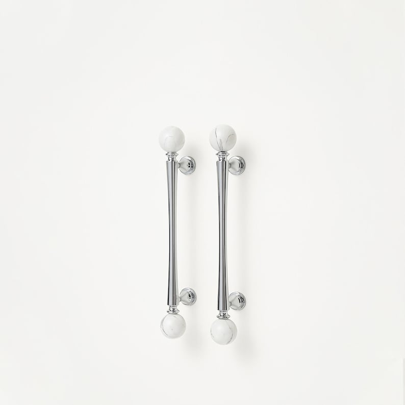 May include: A pair of polished chrome door handles featuring white marble sphere details. The handles have a slender, elongated form with a flared central section. Marble spheres are positioned at the top and bottom of each handle, enhancing the design.