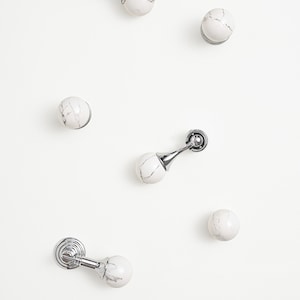 May include: White marble cabinet knobs and door stops with silver-coloured metal hardware. The knobs are round, and the door stops have a curved design. The marble has grey veining.