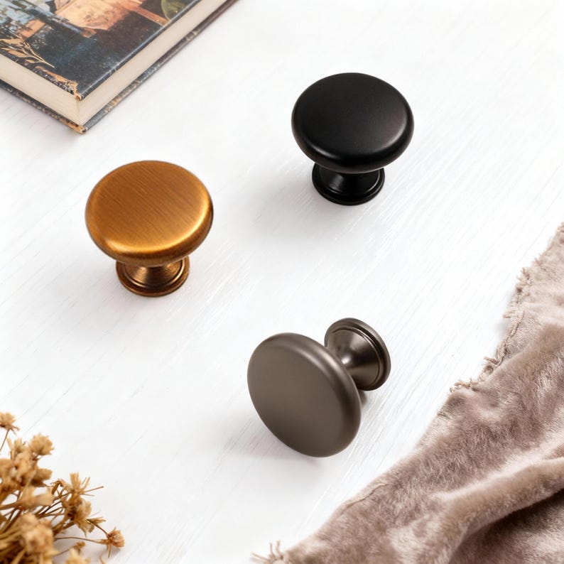 Pu&ograve; includere: Tre pomelli per mobili in diverse finiture: bronzo, nero e grigio. I pomelli hanno una parte superiore rotonda e una base decorativa. I pomelli sono su una superficie bianca con un libro e un tessuto.