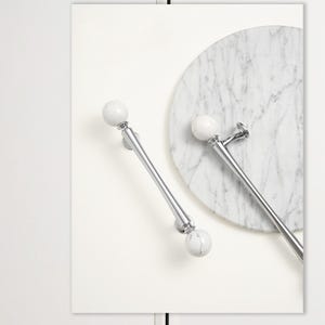 May include: Two polished chrome cabinet pulls with white marble sphere accents. One pull is positioned diagonally, while the other is horizontal. A round marble cutting board is in the background. The pulls are on a white background.