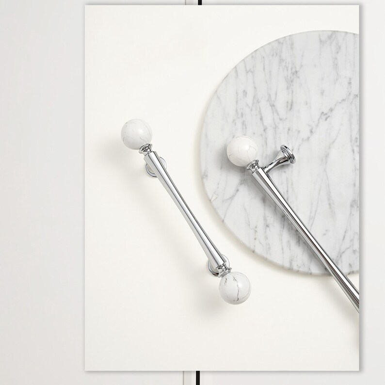 May include: Two polished chrome cabinet pulls with white marble sphere accents. One pull is positioned diagonally, while the other is horizontal. A round marble cutting board is in the background. The pulls are on a white background.