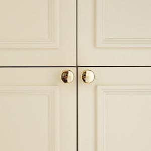 Pu&ograve; includere: Primo piano di quattro ante di armadi color crema con pomelli rotondi dorati. I pomelli sono riflettenti e le ante presentano un design a pannelli incassati. L'immagine &egrave; ben illuminata, evidenziando l'hardware e le linee pulite dei mobili.