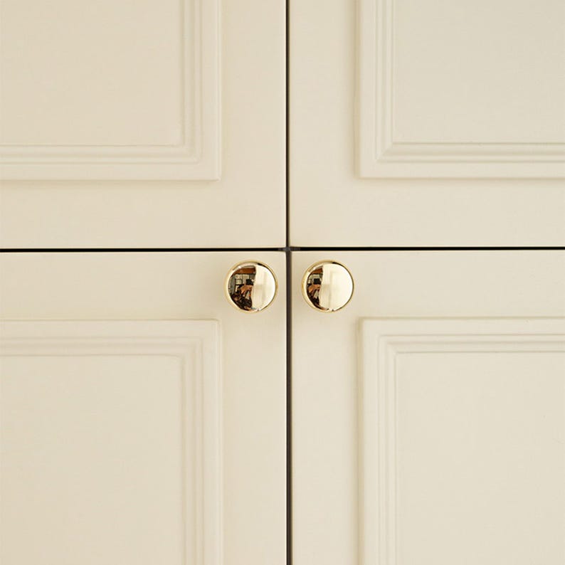 Pu&ograve; includere: Primo piano di quattro ante di armadi color crema con pomelli rotondi dorati. I pomelli sono riflettenti e le ante presentano un design a pannelli incassati. L'immagine &egrave; ben illuminata, evidenziando l'hardware e le linee pulite dei mobili.