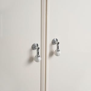 May include: Two cabinet pulls with a polished chrome finish and white marble-effect spherical knobs. The pulls are mounted on a white cabinet door. The design is modern and elegant.