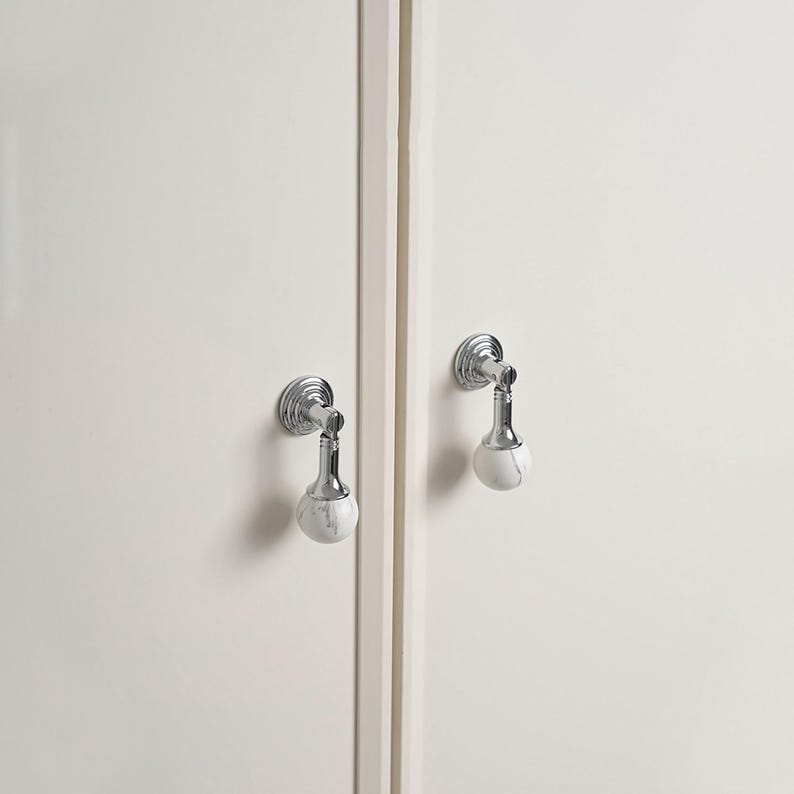May include: Two cabinet pulls with a polished chrome finish and white marble-effect spherical knobs. The pulls are mounted on a white cabinet door. The design is modern and elegant.
