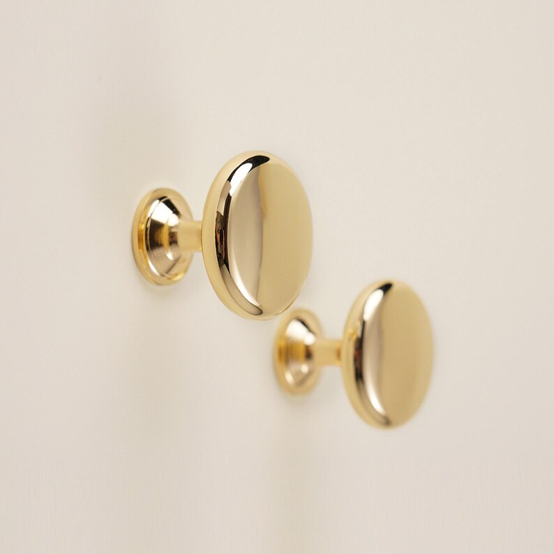 Pu&ograve; includere: Due pomelli per mobili dorati e lucidi con un design ovale. I pomelli sono montati su una parete di colore chiaro, mettendo in risalto la loro superficie riflettente e lo stile semplice ed elegante. Questi elementi hardware sono ideali.