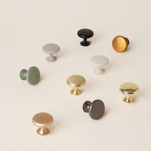 Pu&ograve; includere: Una collezione di pomelli per mobili in vari colori, tra cui oro, nero e verde. I pomelli hanno un design rotondo a forma di fungo e sono realizzati in metallo. I pomelli sono disposti su una superficie di colore chiaro.