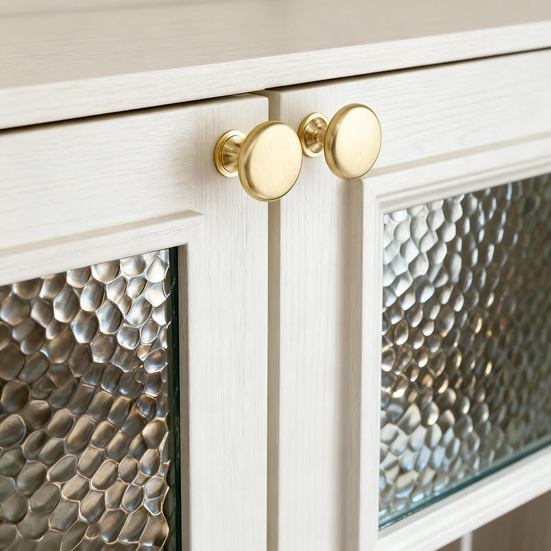 Pu&ograve; includere: Primo piano di un mobile bianco con due ante e pannelli di vetro strutturato. Le ante del mobile hanno pomelli rotondi color oro. I pannelli di vetro hanno una texture unica e a bolle, che aggiunge interesse visivo al design.