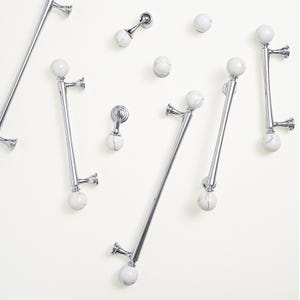 May include: A selection of polished chrome cabinet pulls and knobs, featuring white and grey marble-effect spherical accents. The hardware includes various sizes of bar pulls and round knobs, all mounted on a neutral background.