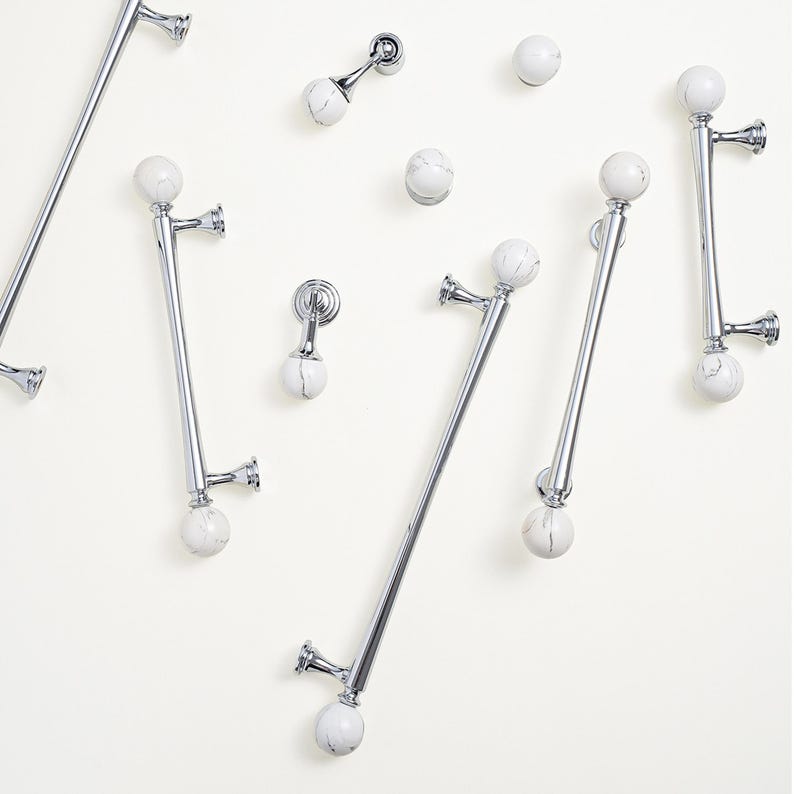 May include: A selection of polished chrome cabinet pulls and knobs, featuring white and grey marble-effect spherical accents. The hardware includes various sizes of bar pulls and round knobs, all mounted on a neutral background.