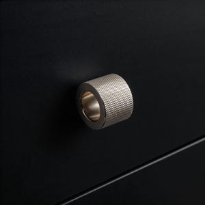 May include: A silver metal cabinet knob with a textured, diamond-patterned surface. The knob is cylindrical with a circular opening and is mounted on a black cabinet door. The hardware is a modern design.