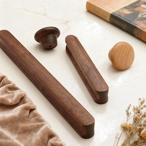 May include: A collection of wooden cabinet hardware, including two long, rectangular pulls, two knobs, and a small, rounded handle. The hardware is made of dark brown wood and sits on a white surface. A book and a velvet bag are also in the image.