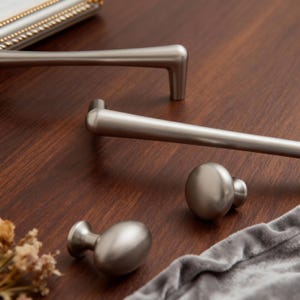 May include: A collection of brushed nickel cabinet hardware, including two bar pulls and two oval knobs. The hardware is displayed on a dark wood surface with a soft gray cloth.