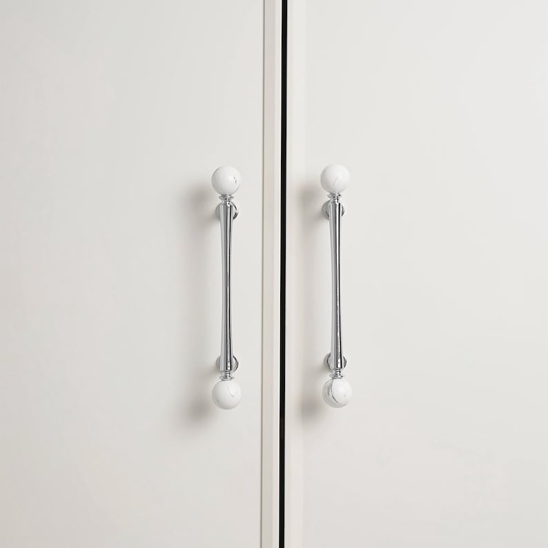 May include: Two white cabinet doors with matching handles. Each door has a polished chrome handle with a white, marble-effect sphere at each end. The handles are vertically aligned, adding elegance to the minimalist design.