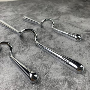 Könnte beinhalten: Drei polierte Werkzeuge aus verchromtem Metall mit einem geschwungenen, hakenartigen Griffdesign. Jedes Werkzeug hat eine glatte, reflektierende Oberfläche und ist in der Nähe des abgerundeten Endes mit dem Text 0452 graviert. Die Objekte liegen diagonal.