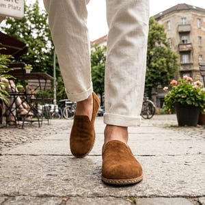 May include: Brown suede slip-on shoes with a rounded toe and a stitched sole. The shoes are worn with white pants, cuffed at the ankle. The image is taken from a low angle, showing the shoes in focus on a cobblestone street.