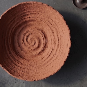 May include: A round, terracotta-colored bowl with a rough, uneven texture. The interior surface features a prominent, hand-formed spiral pattern that radiates from the center toward the jagged, organic-looking rim of the vessel.